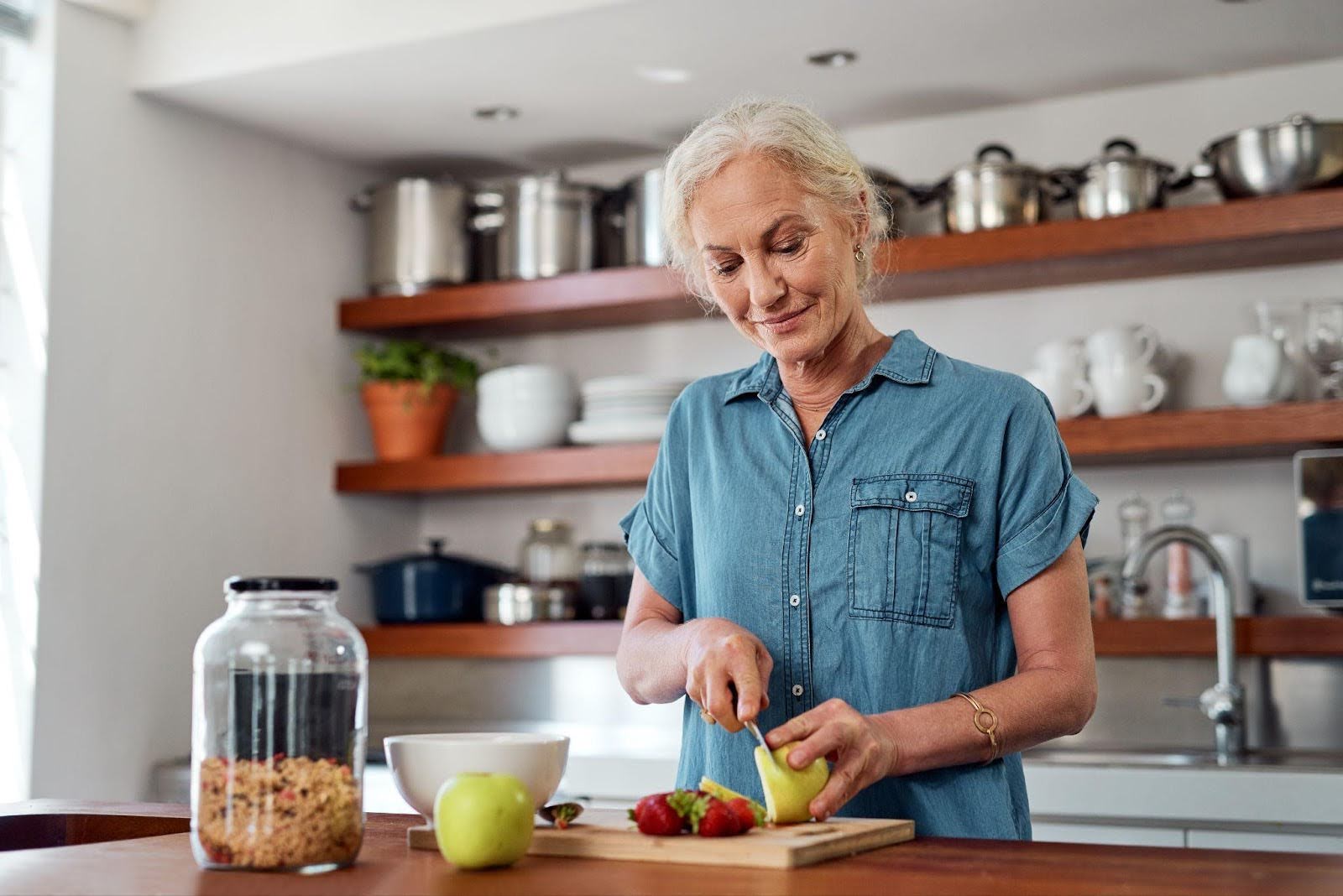 An older woman slices an apple and places the pieces on a wooden cutting board in a bright kitchen