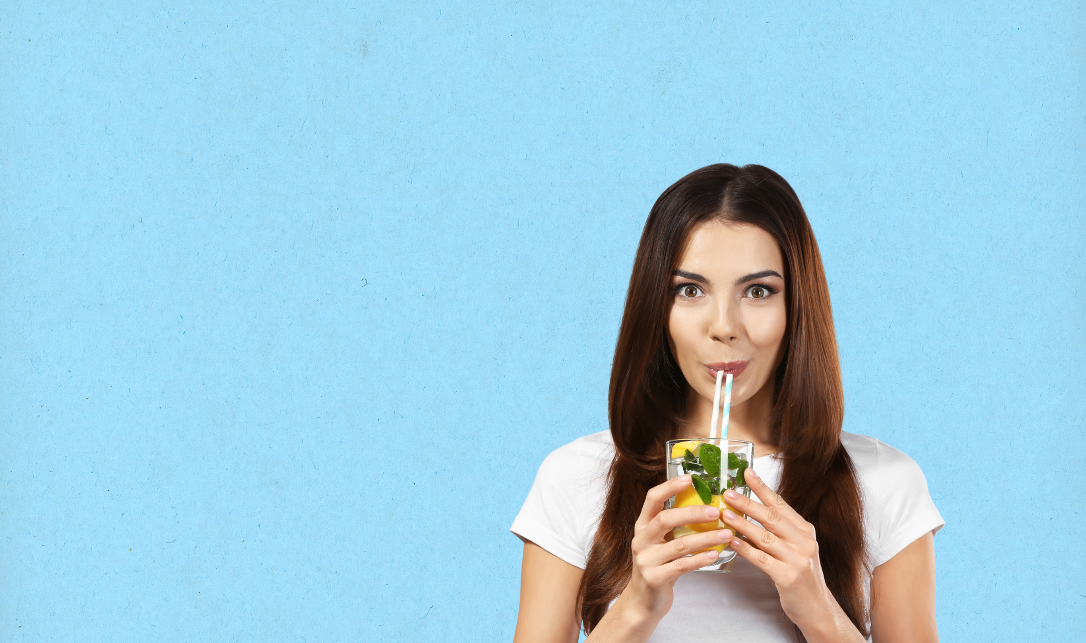 A woman in a white shirt enjoys a refreshing drink with lemon and mint, using a striped straw. She stands against a light blue background, smiling subtly.
