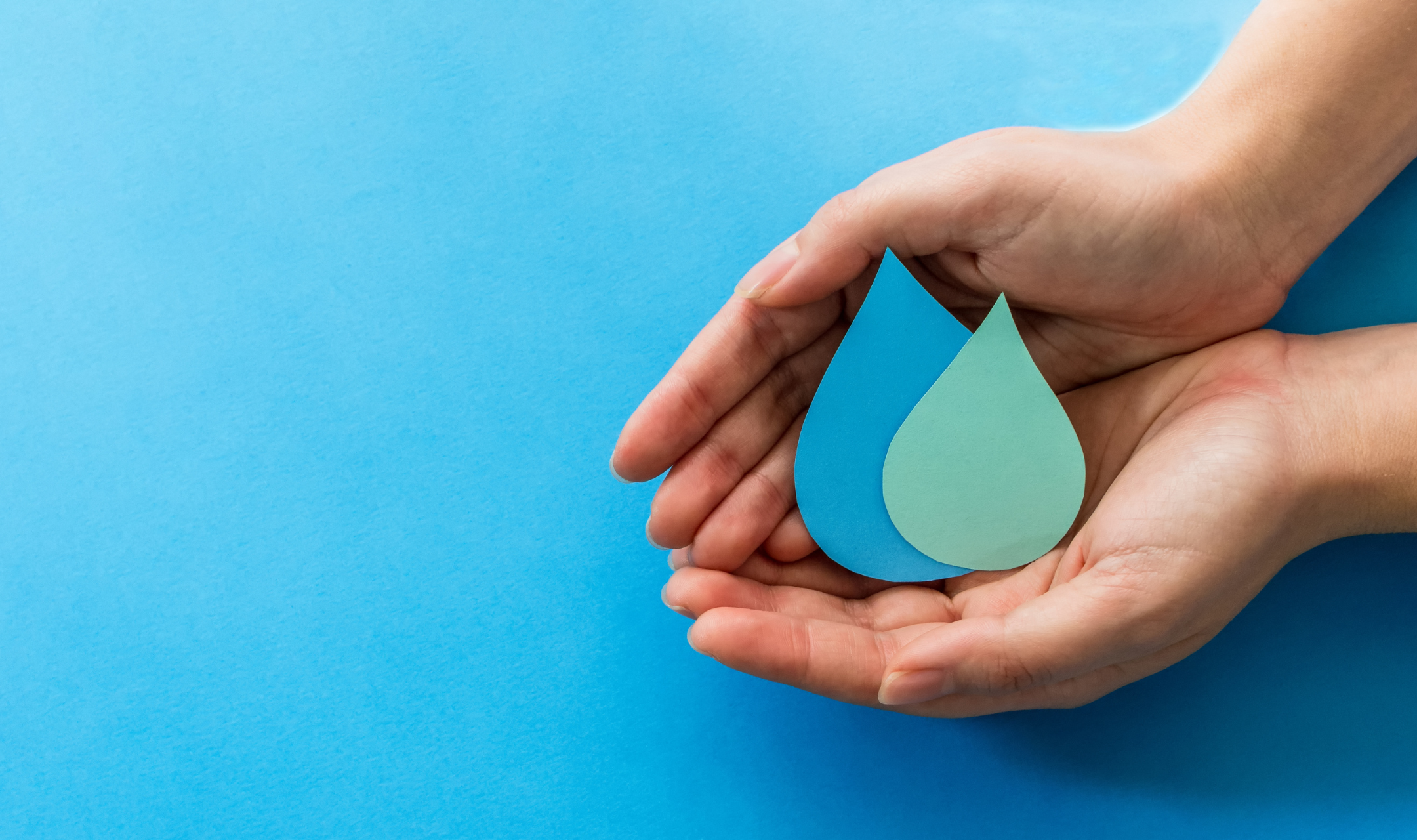 Hands gently cradle two paper water drops, one blue and one teal, against a bright blue background, symbolizing conservation and care.